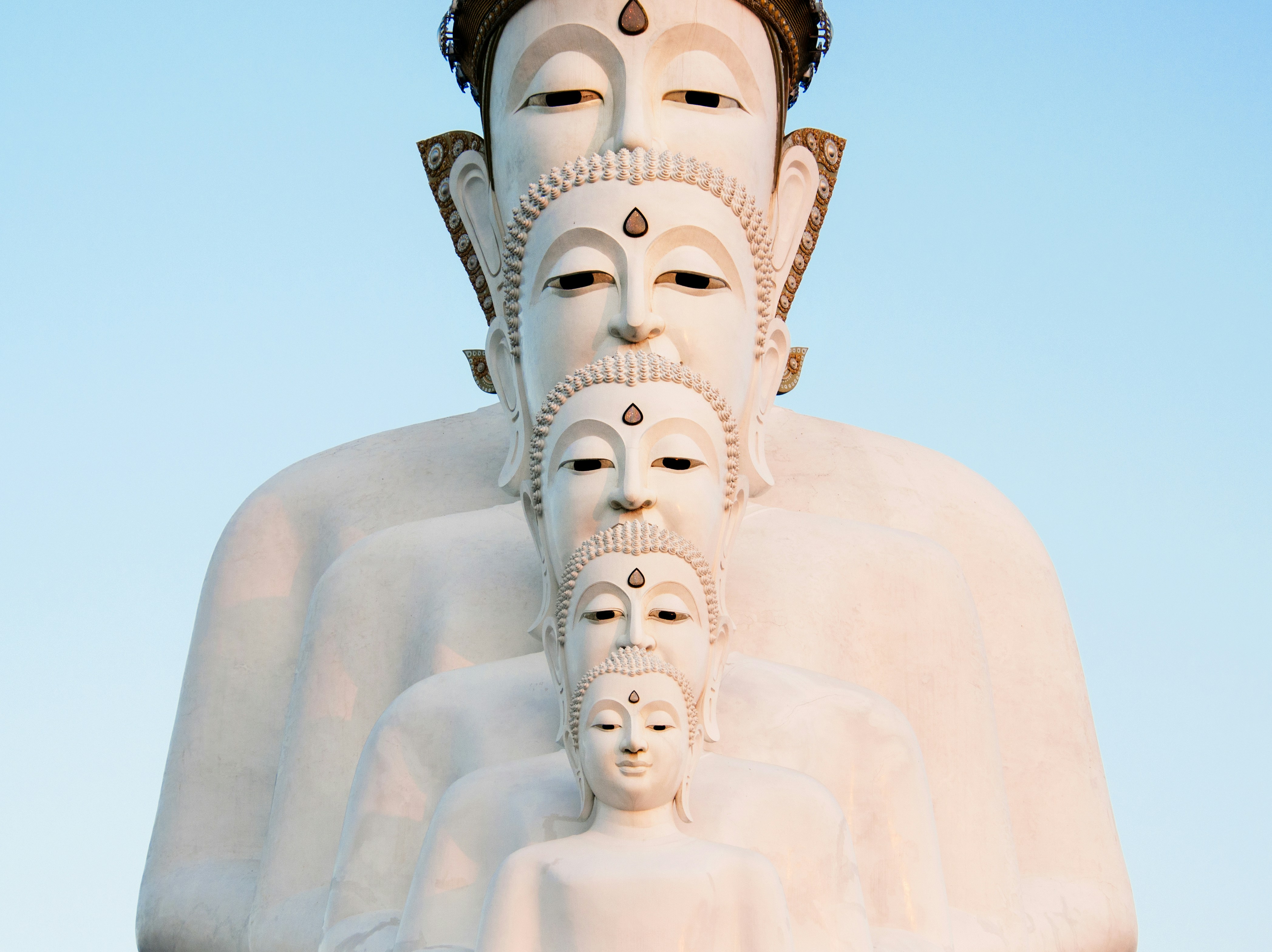 Photograph of five white Buddha statues stacked largest to smallest in symmetrical alignment against a pale blue sky.